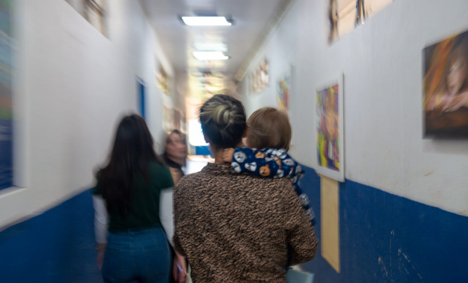 Imagem que mostra um corredor com paredes brancas na parte superior e azuis na parte inferior. Quatro pessoas estão caminhando no corredor, de costas para a câmera. A pessoa mais próxima carrega uma criança pequena em seus braços. As outras três pessoas são adultas, com cabelos longos e roupas casuais. As paredes do corredor estão decoradas com diversos quadros coloridos.