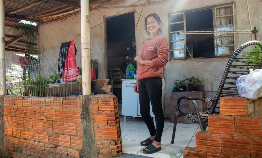 Imagem que mostra a senhora Amélia sorrindo em frente de sua casa. Ela tem cabelos escuros e veste uma blusa de manga comprida cor de telha, calças pretas e chinelos escuros. Ela está em uma área externa coberta, com piso de ladrilho claro. Ao fundo, à esquerda, há uma área mais aberta com vegetação e roupas penduradas em um varal improvisado. A casa é simples, tem paredes rebocadas e uma janela gradeada com cortinas brancas. A luz do sol entra pela lateral direita da imagem, criando um efeito de brilho.