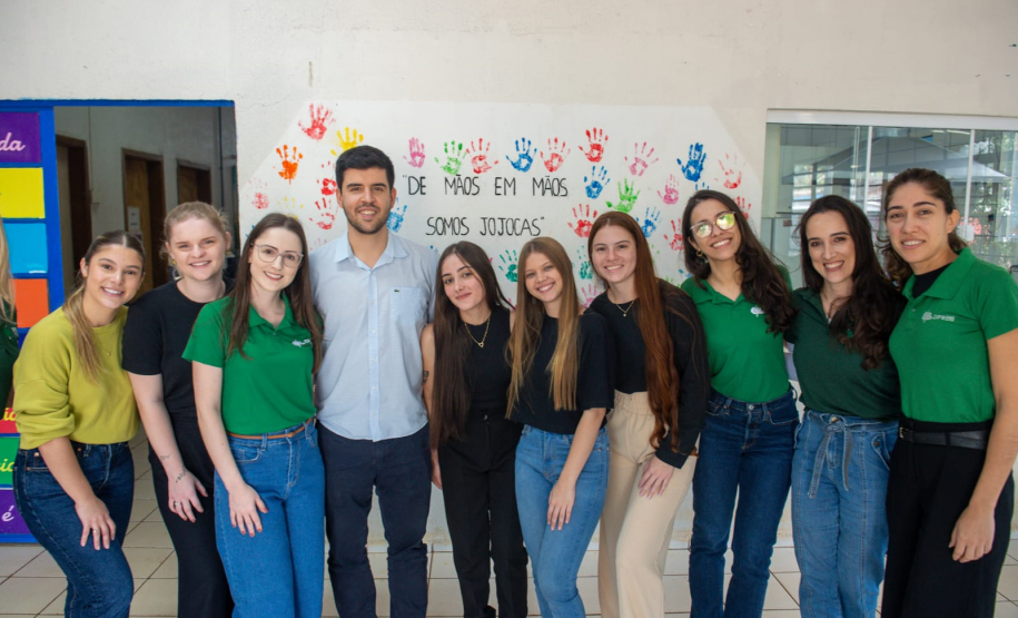 Imagem que mostra a equipe da Defensoria Pública. São nove pessoas, que sorriem posando para a foto em um ambiente interno. Há um homem e oito mulheres, todos com aparência jovem. A maioria das mulheres veste camisetas verdes e calças jeans. O homem veste uma camisa clara de botões e calças escuras. Ao fundo, na parede branca, há diversas impressões de mãos coloridas e a frase "DE MÃOS EM MÃOS SOMOS JOJOCAS" escrita em letras pretas.