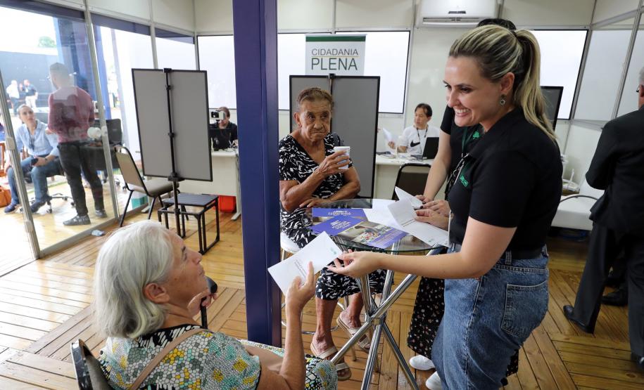 Imagem colorida horizontal em que se vê uma senhora idosa recebendo um panfleto de uma moça com informações sobre os serviços da DPE-PR durante o evento Assembleia Itinerante realizado em Londrina.