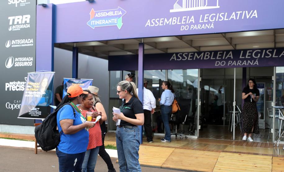 Imagem colorida horizontal em plano aberto em que se vê duas moças recebendo um panfleto de uma moça com informações sobre os serviços da DPE-PR durante o evento Assembleia Itinerante realizado em Londrina.