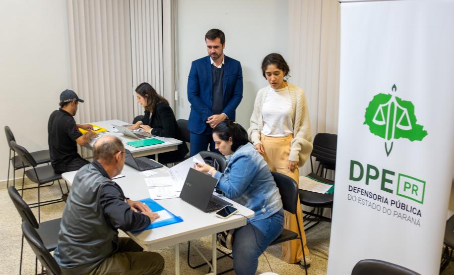 Imagem colorida horizontal em que se vê o banner da DPE-PR em primeiro plano e ao fundo o DPG acompanhando os atendimentos do mutirão.