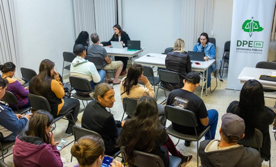 Imagem colorida horizontal em que se vê uma sala cheia de pessoas e ao fundo a equipe da DPE-PR realizando atendimento.