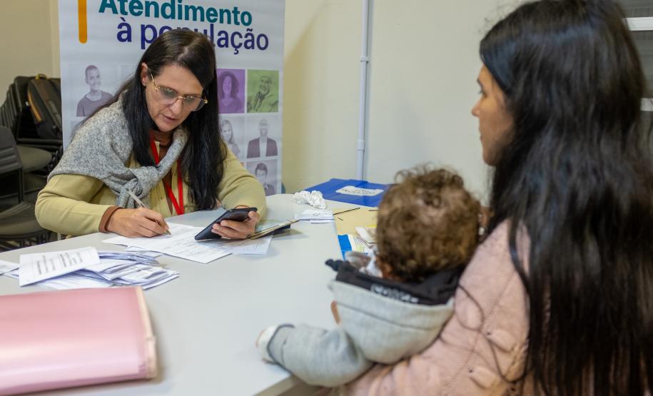 Imagem colorida horizontal em que se vê uma moça de costas segurando um bebê também de costas e ao fundo servidora do MPPR realizando atendimento durante o mutirão conjunto com a DPE-PR.