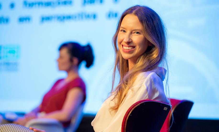 Imagem horizontal colorida de duas mulheres, uma em primeiro plano sorrindo e a segundo ao fundo desfocada.