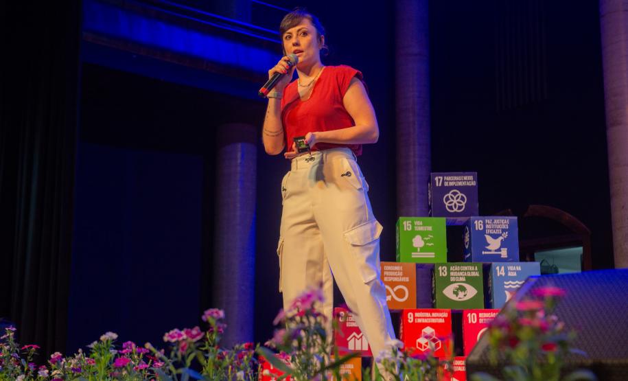 Imagem horizontal colorida de um palco em que se vê a psicóloga DPE-PR falando durante oficina de capacitação sobre o Projeto Ampara.
