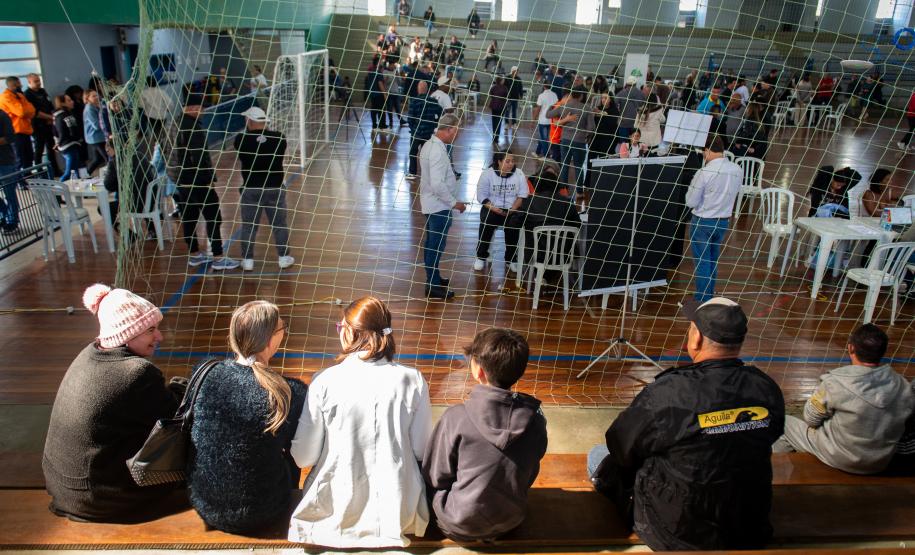 Imagem colorida mostra em plano geral o ginásio com muitas pessoas circulando entre os serviços enquanto alguns aguardam a vez nos bancos do ginásio.
