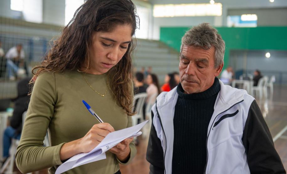Imagem horizontal colorida em que se vê servidora da DPE-PR anotando dados num papel ao lado de um assistido durante Mutirão realizado em Almirante Tamandaré.