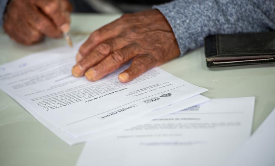 Imagem colorida horizontal que mostra em close as mãos de um homem assinando um documento durante Mutirão da DPE-PR em Almirante Tamandaré.