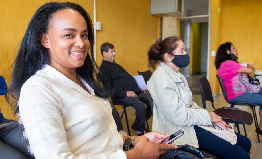 Imagem colorida horizontal em que se vê uma moça negra de cabelos compridos em primeiro plano e ao fundo outros assistidos enquanto aguardam atendimento do mutirão.