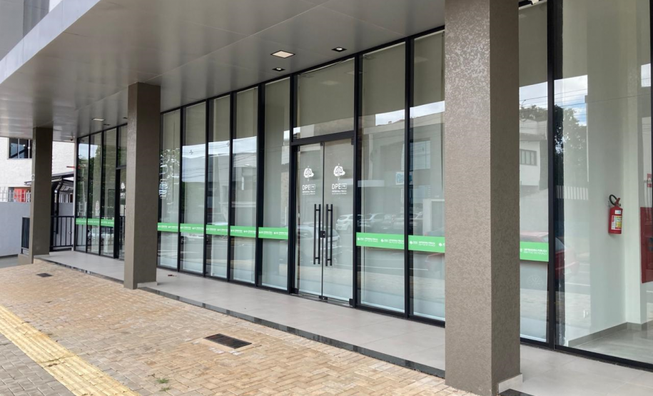 Imagem que mostra a nova sede da Defensoria em Foz do Iguaçu, com uma fachada composta por grandes painéis de vidro. Há pilares cinza-escuro que separam os painéis de vidro e sustentam a estrutura superior do prédio. Na parte inferior dos painéis de vidro, há uma faixa horizontal verde clara com o logo da DPE-PR. Em frente à entrada, há um piso de tijolos que se estende por toda a fachada, com uma faixa amarela tátil para deficientes visuais.