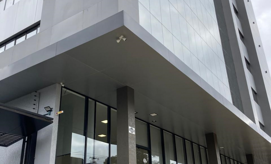 Imagem que mostra a nova sede da Defensoria em Foz do Iguaçu, com uma perspectiva mais ampla da fachada do prédio. O edifício apresenta um design moderno com múltiplas camadas e uma combinação de grandes painéis de vidro espelhados. Uma faixa horizontal verde clara com o logo da DPE-PR percorre a fachada. Uma rampa acessível com piso tátil amarelo leva à entrada principal e há também um corrimão de metal preto. O céu está nublado, o que confere uma iluminação difusa ao ambiente externo.