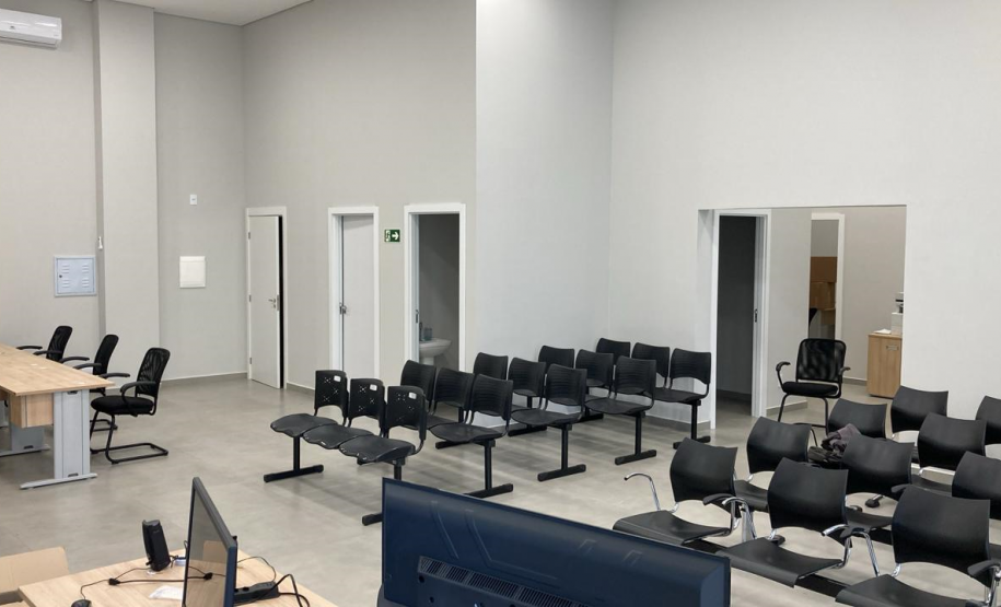 Imagem que mostra a área de atendimento e espera da nova sede da Defensoria em Foz do Iguaçu. O espaço é amplo, com paredes em um tom neutro de cinza claro e piso cinza mais escuro. Há múltiplas fileiras de cadeiras pretas e, no canto esquerdo da imagem, vê-se uma estação de trabalho com mesas de madeira clara e cadeiras de escritório pretas, onde equipamentos eletrônicos como computadores e monitores estão posicionados.