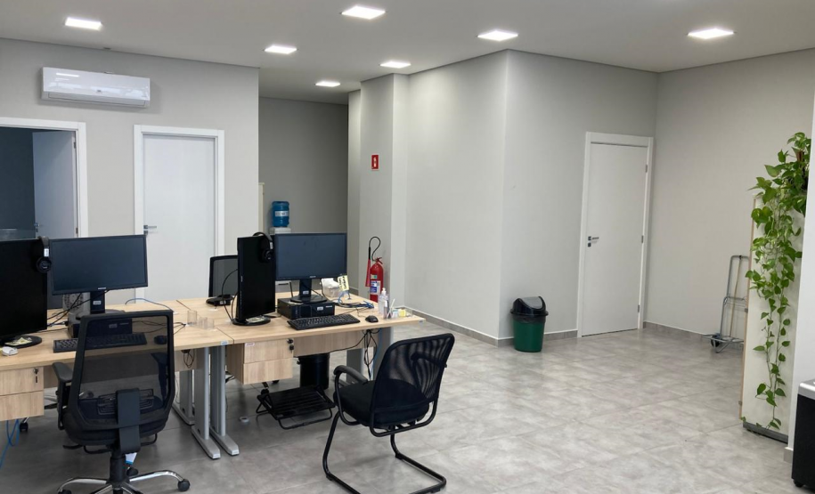 Imagem que mostra uma área de trabalho interna da nova sede da Defensoria em Foz do Iguaçu. O espaço é amplo e bem iluminado por várias luminárias de teto quadradas embutidas. No lado esquerdo da imagem, há duas estações de trabalho com mesas de madeira clara, equipadas com computadores, monitores pretos e cadeiras de escritório. No canto direito da imagem, uma planta “jiboia” com folhas penduradas adiciona um toque de natureza ao ambiente.