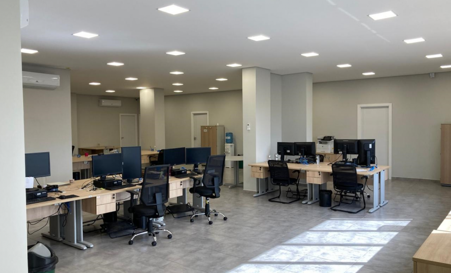 Imagem que mostra a nova sede da Defensoria em Foz do Iguaçu, apresentando uma ampla área de escritórios, com várias estações de trabalho dispostas em um layout de plano aberto. O ambiente é bem iluminado e existem diversas mesas de madeira clara com estruturas metálicas, equipadas com computadores e cadeiras de escritório. Ao fundo, há um bebedouro de água com galão.