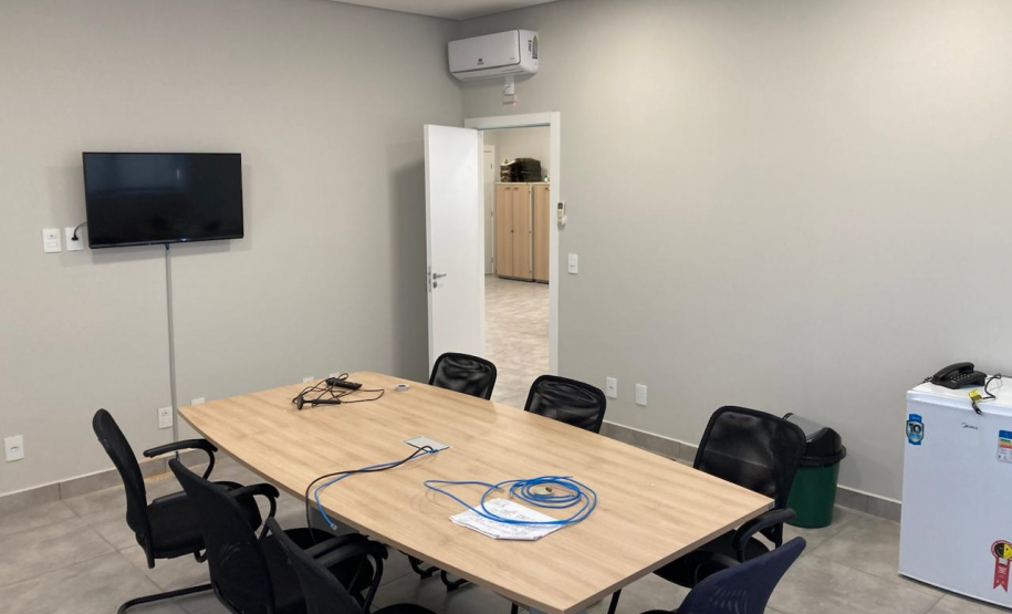 Imagem que mostra uma sala de reunião da nova sede da Defensoria em Foz do Iguaçu. Ela está equipada com uma mesa grande de madeira clara no centro. Ao redor da mesa, há várias cadeiras pretas e em cima da mesa, alguns cabos azuis de conexão à internet e documentos estão visíveis. Em uma das paredes, há uma televisão de tela plana montada e, à direita da imagem, um frigobar branco.