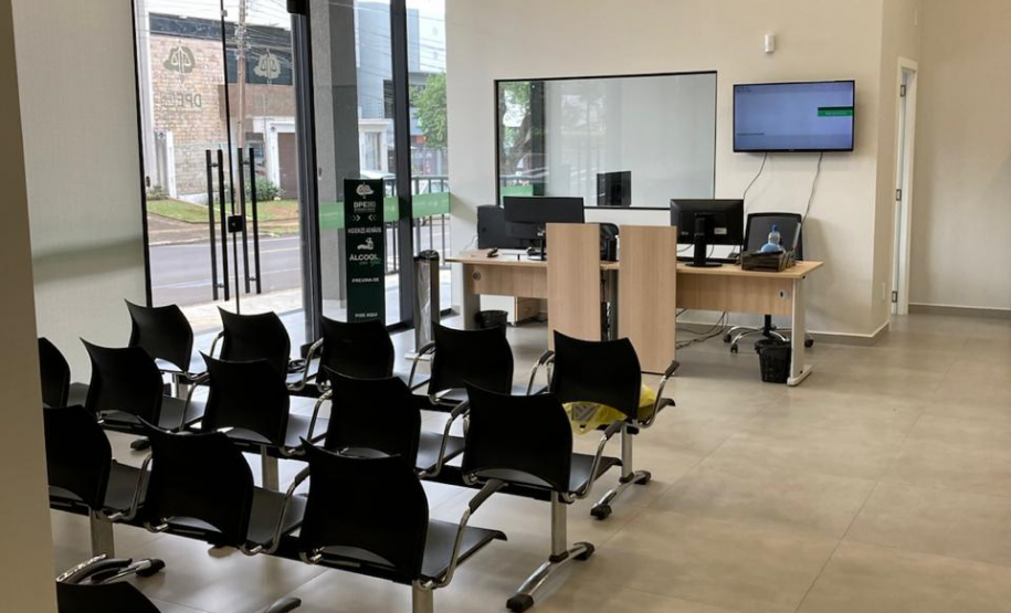 Imagem que mostra a área de recepção da nova sede da Defensoria em Foz do Iguaçu. À esquerda, grandes portas de vidro permitem a entrada de luz natural e oferecem vista para o exterior, onde é possível ver uma rua e vegetação. O ambiente possui fileiras de cadeiras pretas com estrutura metálica, voltadas para uma área de atendimento. Ao fundo, há mesas de madeira clara com computadores e monitores pretos, indicando estações de trabalho para atendimento ao público.