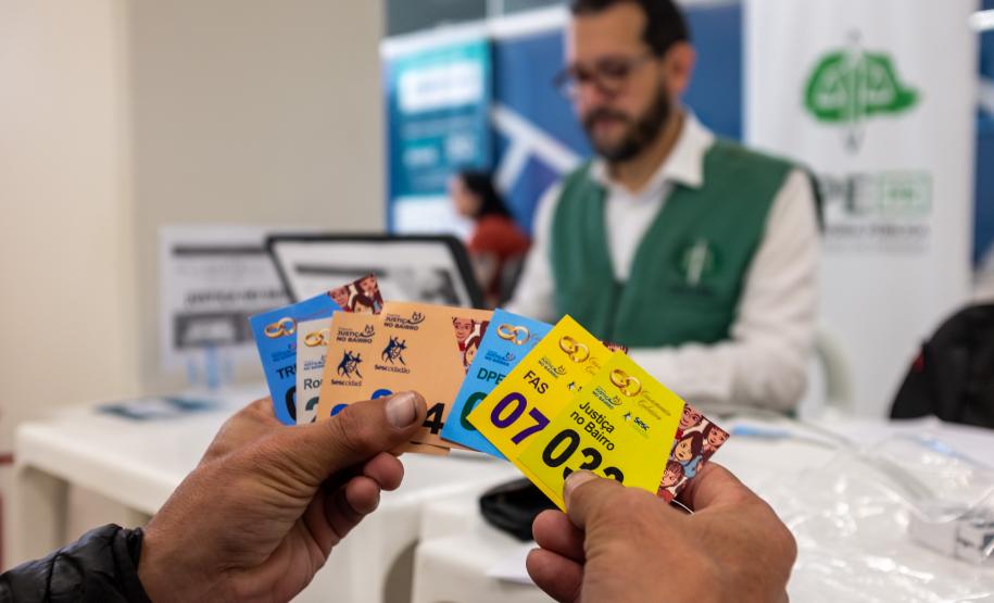 Em primeiro plano se vê as mãos de um assistido segurando as senhas de diversos serviços oferecidos no Registre-se e ao fundo desfocado o defensor público que fazia o seu atendimento.