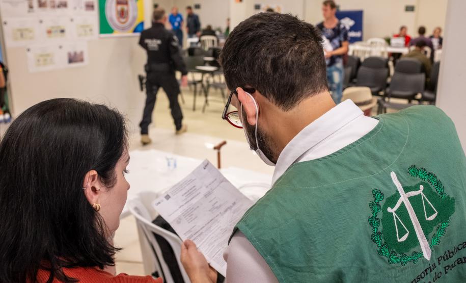 Imagem em que se vê Defensor Público de costas segurando os documentos de um usuário. Ao fundo é possível ver os guichês de atendimento do Registre-se.