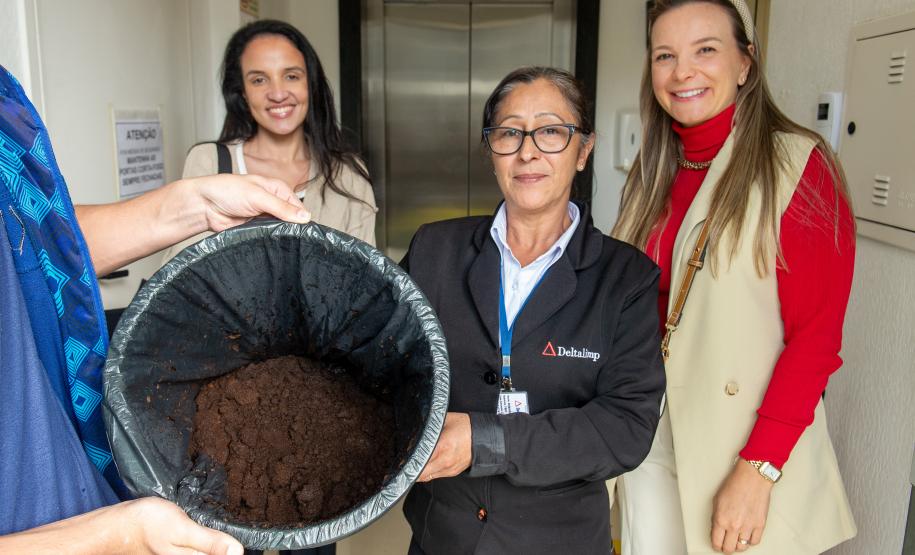 Três mulheres posam com a borra de café na sede da Defensoria. Toda borra do café passado na instituição será entregue no Ecoponto da Prefeitura de Curitiba onde vira adubo em um minhocário.