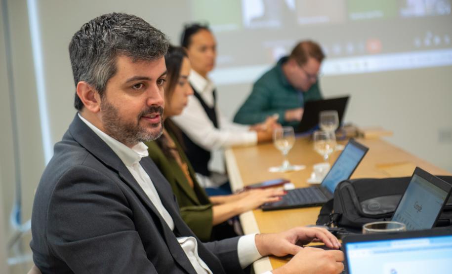 Em primeiro plano se vê homem de cabelo levemente grisalho e terno e ao fundos outras duas pessoas