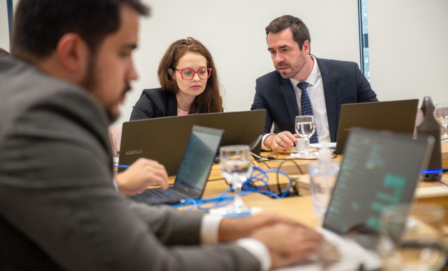 Imagem de um homem de terno em primeiro plano e ao fundo um homem de terno e uma mulher de óculos vermelho conversam olhado a tela de um laptop.