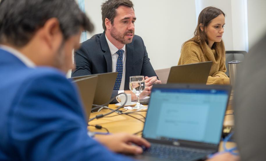 A imagem mostra pessoa de costas em primeiro plano e ao fundo se vê um homem e uma mulher; todos estão usando laptops.