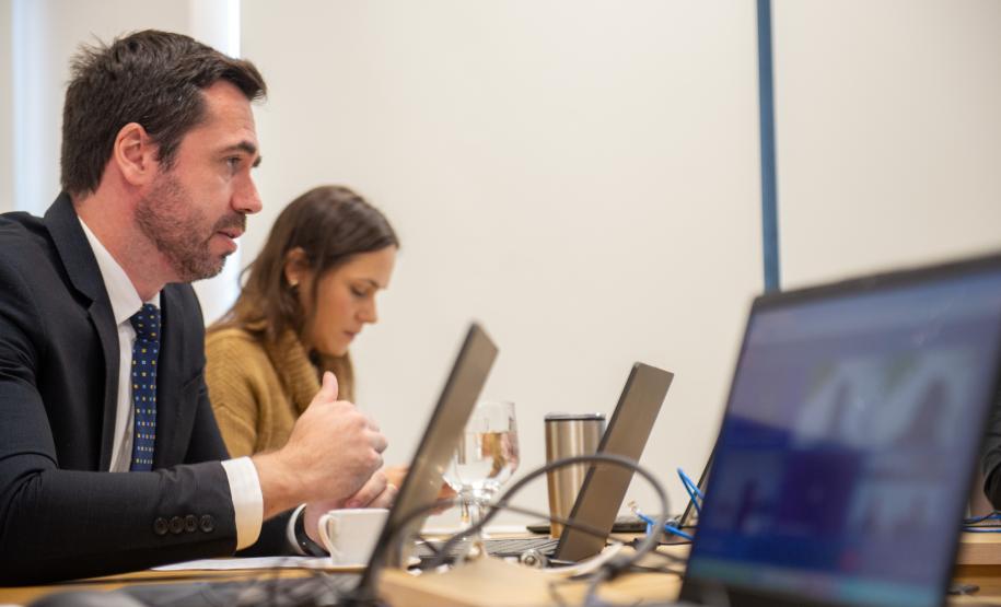 Um homem de terno e uma mulher acompanham reunião ao computador e em primeiro plano tela de laptop em que aparecem os participantes online da reunião.