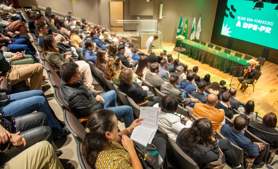 Imagem mostra o auditório quase cheio para a posse dos servidores, no palco ocorre uma apresentação músicas de violão e voz. Ao fundo mesa para as autoridades e o telão com logo da DPE-PR.