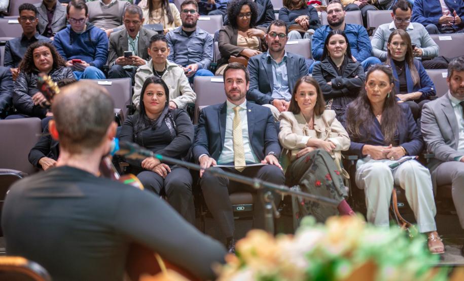 Imagem onde se vê músico tocando violão de costas e ao fundo as cadeiras lotadas do auditório.