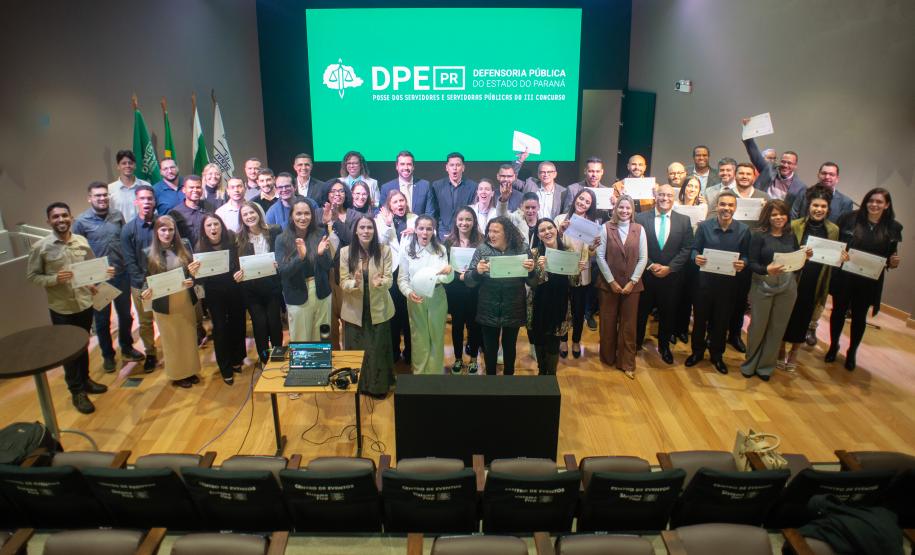 Grupo de Servidores empossados posam juntos para foto no palco de auditório. Ao fundo tela de Led mostra a logo da instituição.
