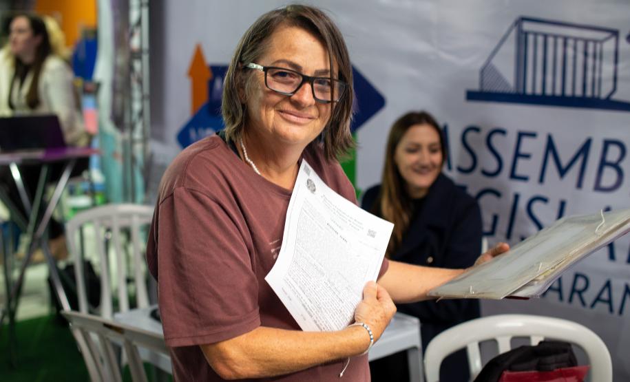 Mulher assistida pela DE-PR posa para a foto com os documentos ao fim do  atendimento ontem no ALEP nos Bairros.