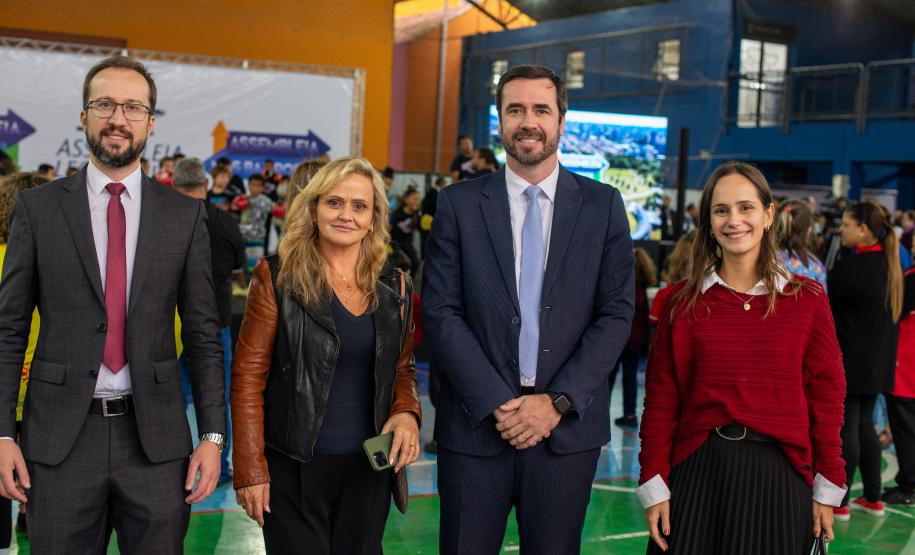 Quatro pessoas posam para foto, dois homens de terno e duas mulheres. Ao fundo se vê grande número de pessoas circulando pelo evento.