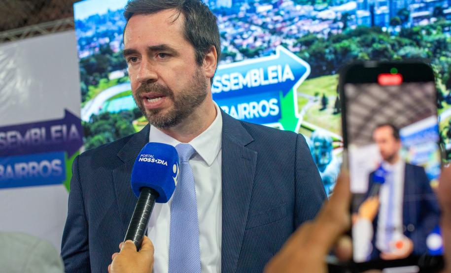 O DPG Matheus Munhoz dá entrevista. Homem de terno fala ao microfone, ao fundo tela de celular grava a entrevista.