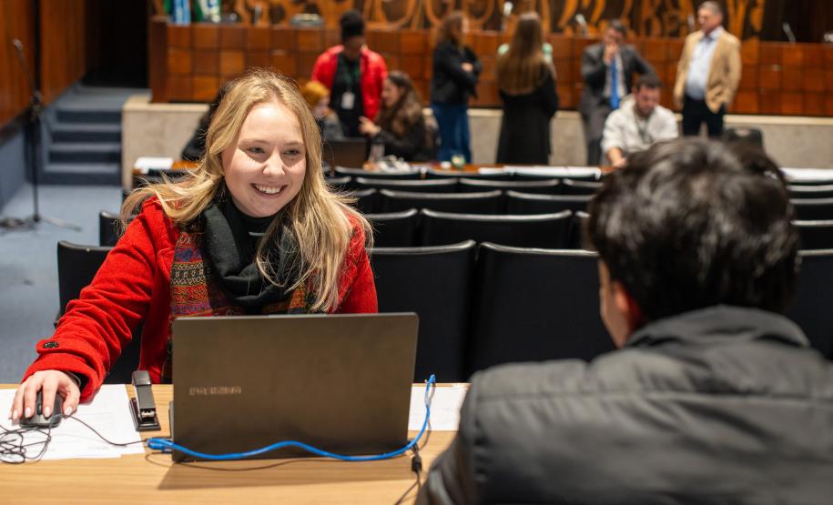 No primeiro plano um homem de costas, ao fundo uma mulher de casaco vermelho realiza atendimento. Ao fundo grupo de pessoas executam outras atividades.