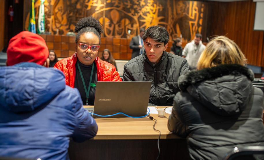 Quatro pessoas sentadas à mesa, dois homens e duas mulheres. Conversam e há um computador na mesa. Ao fundo um painel de madeira do Plenarinho da Assembleia Legislativa.