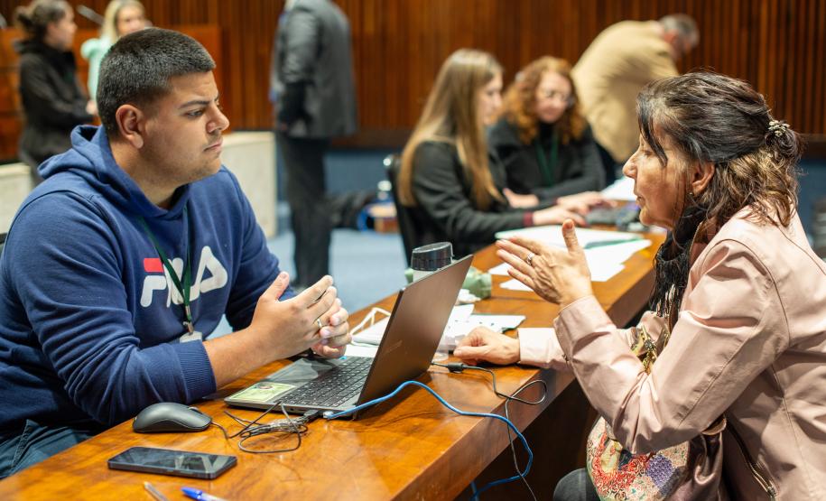 Homem e mulher sentados conversam no primeiro plano. Ao fundo é possível ver outras pessoas trabalhando.