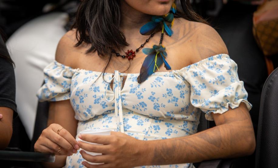 Imagem em detalhe de uma mulher indígena onde se vê o vestido, os braços com pintura corporal e brinco de penas.