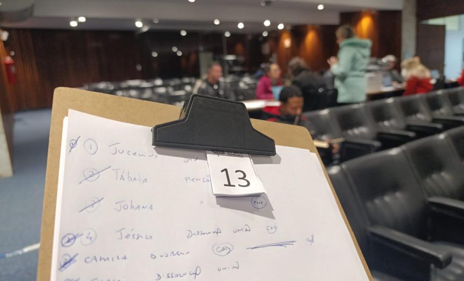 Imagem mostra prancheta com os nomes dos assistidos anotados na triagem e senha em primeiro plano. Ao fundo as pessoas são atendidas.