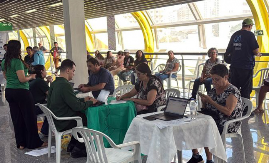 Imagem mostra um grande grupo de pessoas, algumas em pé, outras sentadas à mesa, em mutirão da Defensoria realizado em terminal de ônibus.