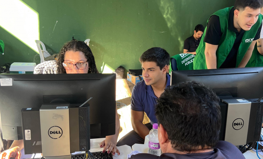 Imagem mostra três servidores utilizando computadores durante os atendimentos do mutirão. De costas em primeiro plano se vê um assistido.