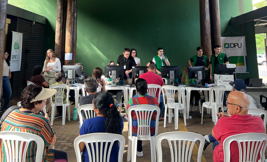 Imagem mostra grupo de pessoas sentadas em cadeiras de plástico no primeiro plano e ao fundo se vê pessoas sendo atendidas em mesas por defensores e servidores da DPE-PR.