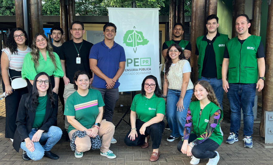 Equipe da DPE-PR posa para foto junto ao Banner da instituição. São 13 pessoas entre homens e mulheres, parte usando coletes verdes da instituição.