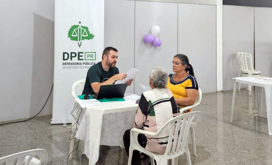Imagem mostra duas mulheres, um idosa, sentadas à mesa conversando com homem jovem usando o computador, Ao fundo se vê banner da Defensoria com a logo da instituição.
