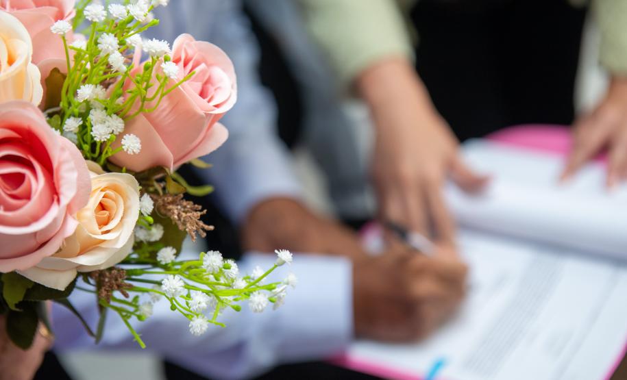 Flores do buquê em primeiro plano. Ao fundo em desfoque um casal assina o documento do cartório.