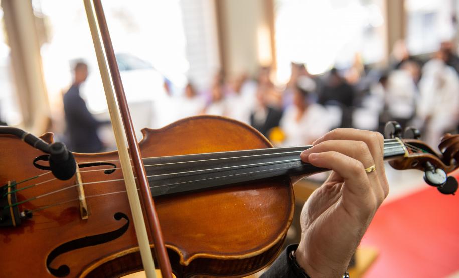 Violino sendo tocado em close-up. Ao fundo aparecem os casais e familiares em desfoque.