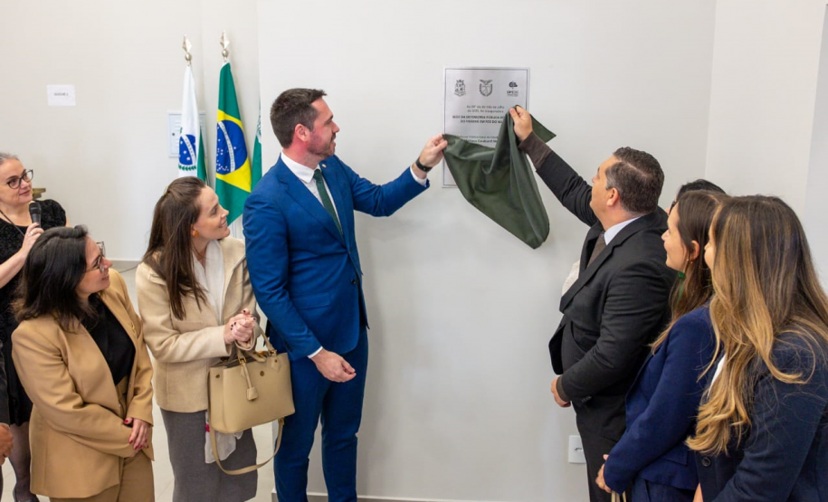 A imagem captura o momento do descerramento da placa inaugural. O Defensor Público-Geral, à esquerda, e o vice-prefeito de Foz do Iguaçu, à direita, seguram juntos um tecido verde-escuro, revelando a placa fixada na parede. Ao redor deles, um grupo de convidados, majoritariamente mulheres, observa a cena com sorrisos e expressões de satisfação.
