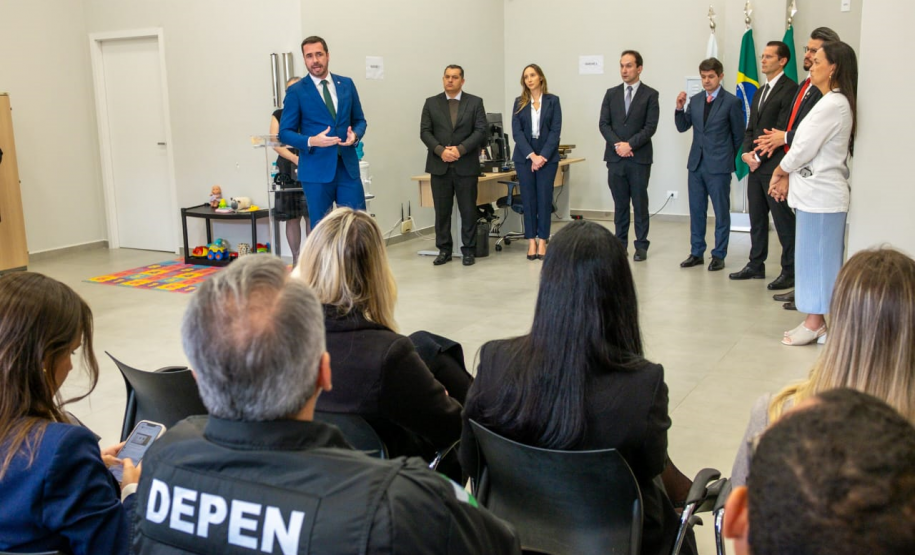 Nesta foto, o Defensor Público-Geral discursa de frente para a câmera. Em pé, logo atrás dele, um grupo de autoridades acompanha o pronunciamento. Em primeiro plano, vemos a plateia sentada de costas, assistindo à cerimônia. Um detalhe notável ao fundo, no canto esquerdo, é um pequeno espaço infantil com tapete colorido e brinquedos, sugerindo o caráter acolhedor do novo local.
