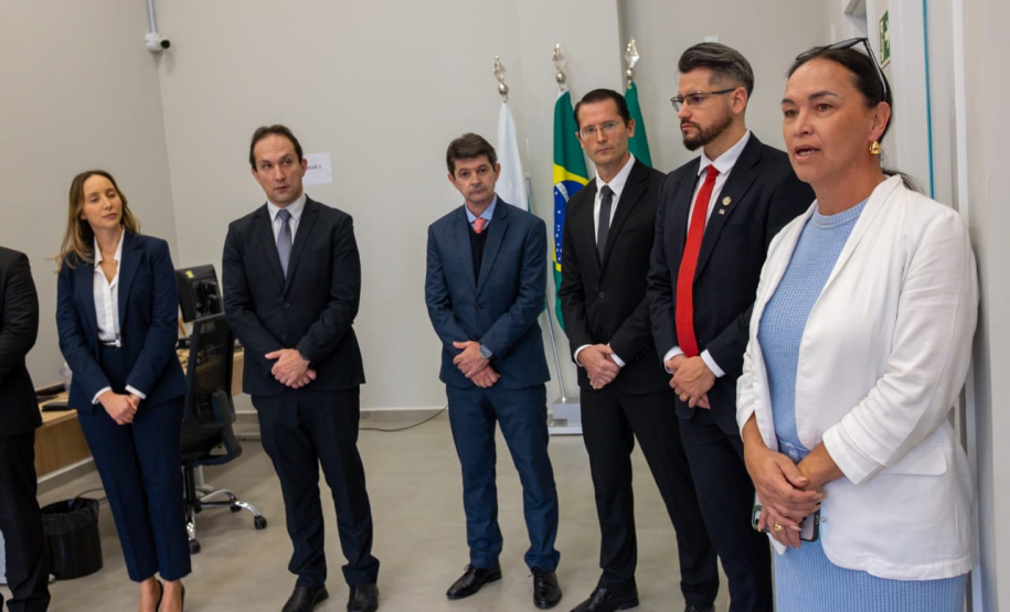 Em primeiro plano, no lado direito da imagem, a Ouvidora-Geral da DPE-PR faz seu discurso. Ela tem uma expressão séria e mantém as mãos unidas à frente do corpo. Ao seu lado, um grupo de autoridades, composto por quatro homens e uma mulher, ouve atentamente. Ao fundo, as bandeiras do Brasil e do Paraná estão hasteadas, compondo o cenário oficial.