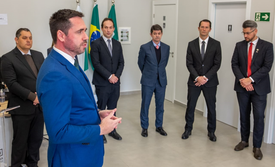 A foto foca no Defensor Público-Geral durante seu discurso, visto em primeiro plano. Com o rosto concentrado, ele gesticula e se dirige a um grupo de cinco homens que o ouvem atentamente. Os homens estão em pé, formando um semicírculo. O ambiente é uma sala ampla com paredes claras e boa iluminação.